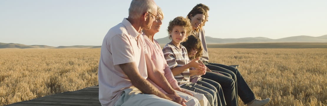 Multigenerational family sitting on a wooden platform at the edge of a golden wheat field, with mountains visible in the distance under a clear sky.